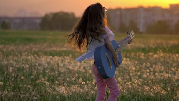 一个美丽的小女孩在夕阳下在蒲公英地里弹吉他 户外远离城市建筑的情感和滑稽的鬼脸 — 图库视频影像