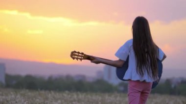 gün batımında güzel manzaralı. komik küçük kız müzisyen gitar çalmak uzak şehir binaları dandelions alanında açık havada yürür. Sanat ve yaratıcılık kavramı