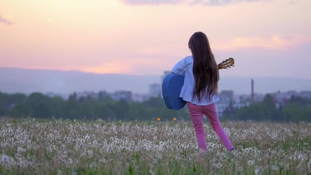 サングラスの都市景観の面白い少女ミュージシャンの背景にタンポポの草原の夕暮れギターと散歩を演奏します コンセプト アートと想像力 — ストック動画