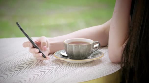 スマート フォンを手に保持している白人少女の体部分手 コーヒー テラスのテーブルでお茶を一杯 — ストック動画