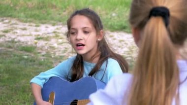 çekici küçük kız akustik gitar çalar ve parkta onun kız arkadaşı ayakta dinleme müzik sanatçı şarkı söylüyor.