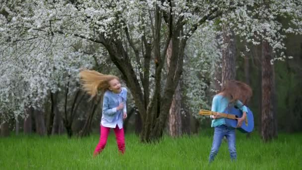 つの学校のガール フレンドは ジャンプとギターを手に笑い浮気春フォレストのロック パフォーマンスを上演しました ツリーの緑の草の純粋な自然の休日とレクリエーションを開花 — ストック動画