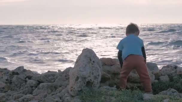 小さな男の子の機能不全の家族は海岸で時間を過ごす 孤児の子供の古いみすぼらしい服 単独で岩ビーチ風の天気をスロー再生します 孤独と社会的不公平を — ストック動画