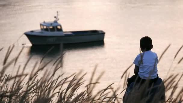 父と息子は一緒に海辺の夏の夕日に向かって歩いています 彼の父は一緒に海岸近くに係留ボートを見て楽しんでいる肩に座って小さな男の子 — ストック動画