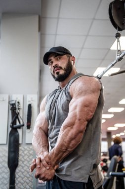 Sakallı, şapkalı ve spor kıyafetli yakışıklı genç adam spor salonunda güçlü kaslarını gösteriyor.