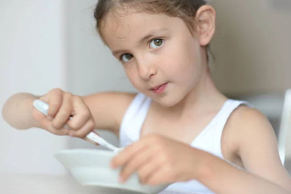 Fată frumoasă caucaziană în rezervor alb de top mâncând micul dejun ținând farfurie și lingură așezată în bucătărie luminoasă — Fotografie, imagine de stoc