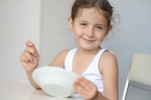 Fericit zâmbind frumoasă fată caucaziană în rezervor alb de sus care deține lingură și farfurie în sala de mese de bucătărie luminoasă — Fotografie, imagine de stoc