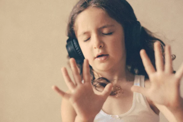 Прелестная маленькая кудрявая девочка с закрытыми глазами в наушниках, слушающая музыку жестом руки — стоковое фото