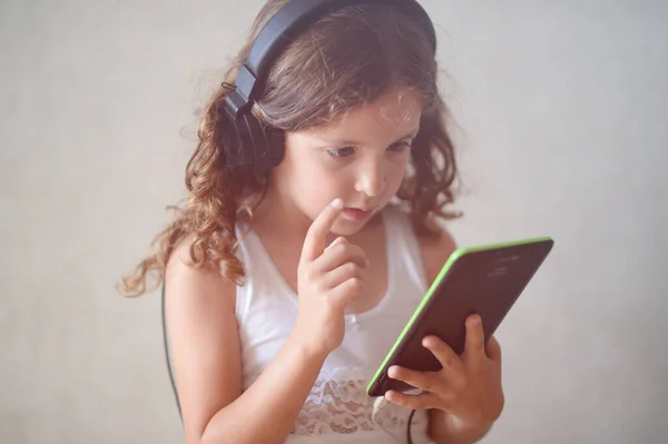 Задумчивая кавказская маленькая кудрявая девочка в наушниках с черным планшетником гаджет онлайн — стоковое фото