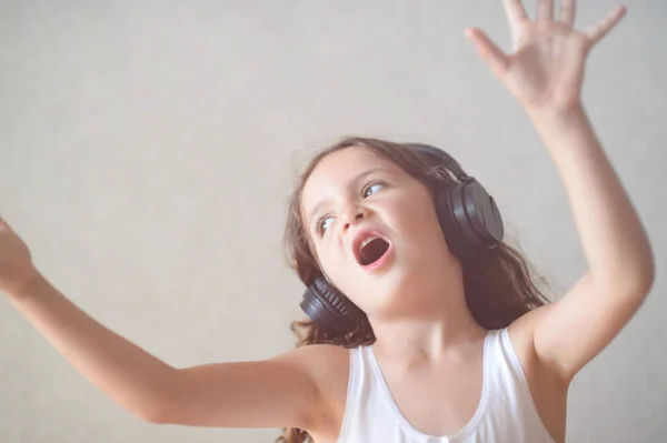Возбужденный маленький белый симпатичная маленькая девочка в черных наушниках поет песню размахивая руками в помещении — стоковое фото