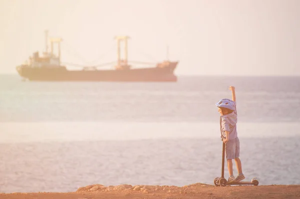 Ευτυχισμένο μικρό παιδί σε κράνος με σκούτερ στη θάλασσα παραλία με φορτηγό πλοίο στον ορίζοντα κατά το ηλιοβασίλεμα — Φωτογραφία Αρχείου