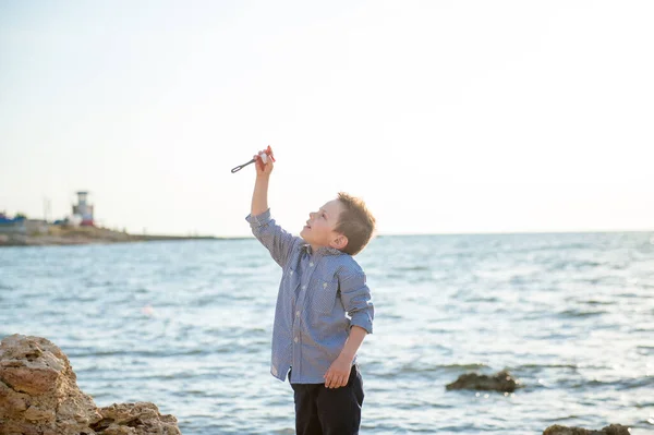 Ενεργό άθλημα μικρό παιδί κρατώντας χαρταετό στη βραχώδη ακτή της θάλασσας κατά το καλοκαιρινό ηλιοβασίλεμα — Φωτογραφία Αρχείου