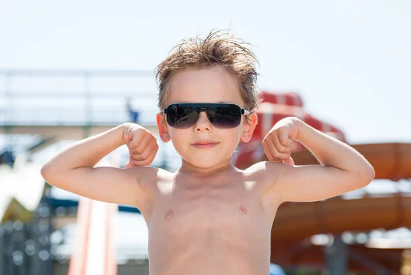 Gezond klein caucasianboy dragen zonnebril tonen sterke spieren in de zomer amusement waterpark — Stockfoto
