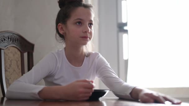 Una pequeña chica caucásica comiendo de plato usando cuchara en casa cocina brillante por la mañana — Vídeos de Stock