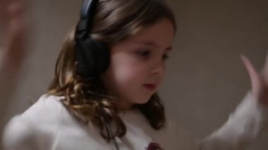 Komik, aktif, küçük, kıvırcık, kulaklıklı beyaz kız evde dans eden müzik dinliyor.