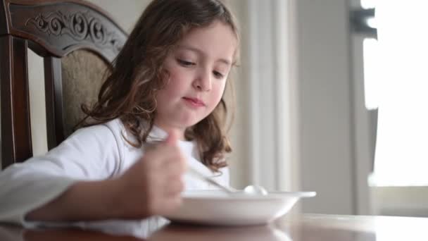 Милая маленькая кудрявая кавказская девочка завтракает молоком в светлой домашней кухне — стоковое видео
