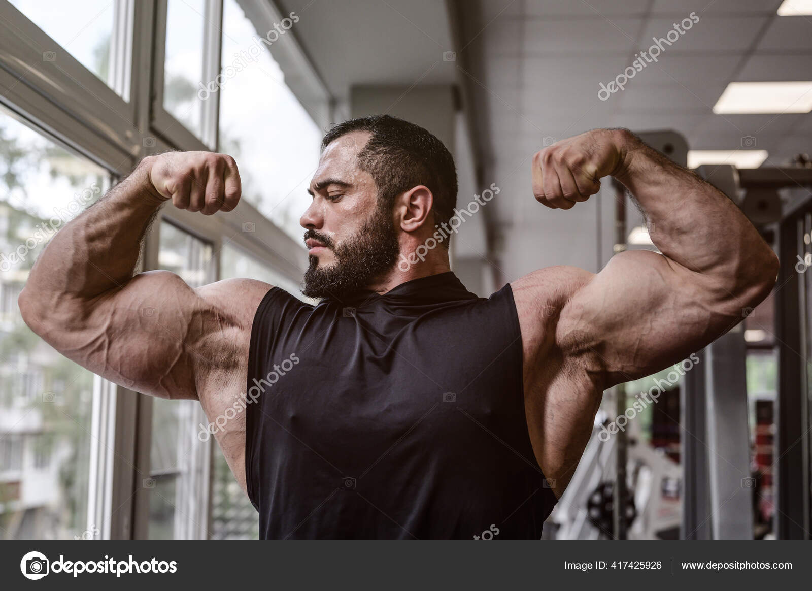 Forte jovem atleta homem com barba vestindo top tanque preto mostrando  grande músculo duplo bíceps no ginásio esporte com janela fotos, imagens de  © photo_zaur.mail.ru #417425926