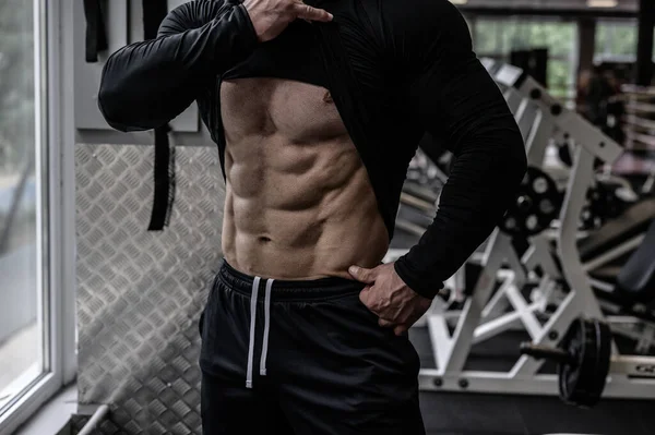 Perfektní tělo s břišními svaly ukazuje mladý silný sportovec poblíž okna ve sportovní tělocvičně s cvičební výbavou — Stock fotografie