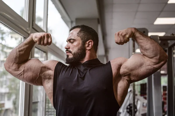 Fuerte joven atleta hombre con barba vistiendo camiseta negra que muestra un gran músculo bíceps doble en el gimnasio deportivo con ventana — Foto de Stock