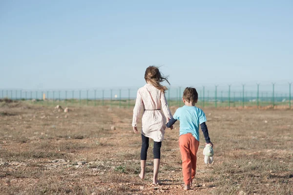 Δύο φτωχά παιδιά οικογενειακός αδελφός με παιχνίδι και λεπτή αδελφή πρόσφυγας λαθρομετανάστης περπατώντας ξυπόλητος μέσα από καυτή έρημο προς τα σύνορα της πολιτείας με συρματόπλεγμα — Φωτογραφία Αρχείου