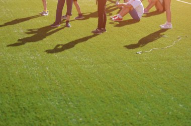 Bir grup çocuk açık havada spor yaparken yeşil futbol sahasında oturup duruyorlar. Okulda boş zamanlarında fotokopi alanı olan bir spor aktivitesi.