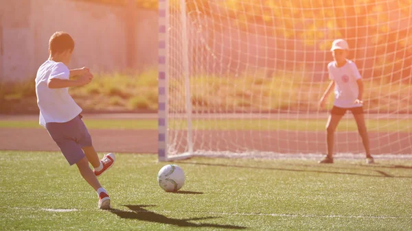 Pequeno garoto ativo chutando bola para o portão de futebol com menino goleiro no campo verde de futebol — Fotografia de Stock