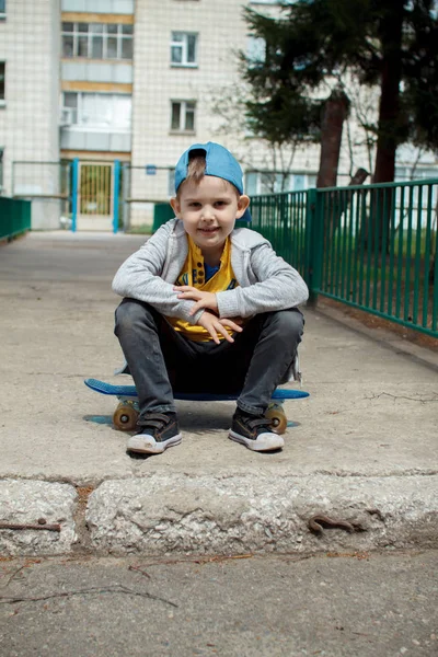 Маленький городской мальчик со скейтбордом. Молодой парень в кепке сидит. Городской стиль. Городские дети. Малыш веселится. — стоковое фото