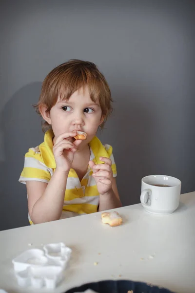 The child sits at a white table by the gray wall, drinking tea and biting cookies. Pensive look in that direction. — Stock Photo, Image