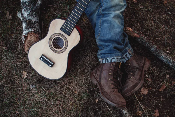 Zbliżenie gitary i stóp w butach. Mały chłopiec w przyrodzie z gitarą. — Zdjęcie stockowe