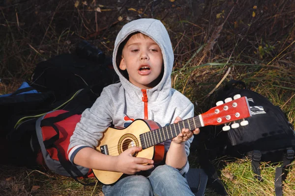 Un niño pequeño con una guitarra se sienta en el parque en la hierba y canta canciones conmovedoras. Niño talentoso Fotos de stock libres de derechos