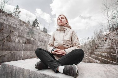 Mermer ocağının arka planında meditasyon eden bir sarışının portresi. Doğa kavramı ile birlik. Rüzgarlıkta bir taşın üzerinde oturan kız.