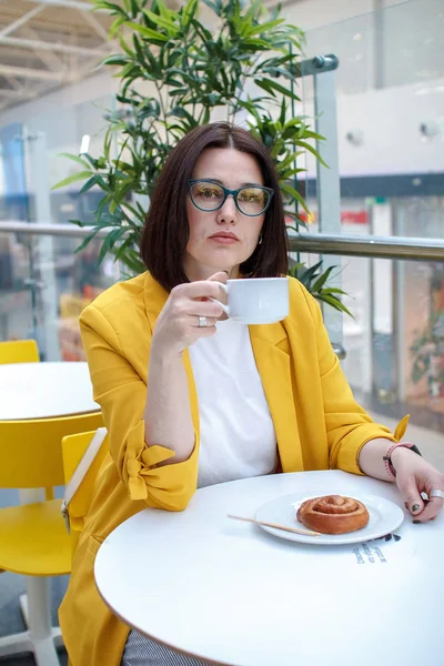 Koffiepauze. Brunette meisje in een gele jas met bril, koffie drinken aan een witte tafel. Rusten na het winkelen. — Stockfoto