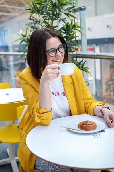 Διάλειμμα για καφέ. Μελαχρινή κορίτσι με ένα κίτρινο μπουφάν με γυαλιά, πίνοντας καφέ σε ένα λευκό τραπέζι. Ανάπαυση μετά τα ψώνια. — Φωτογραφία Αρχείου