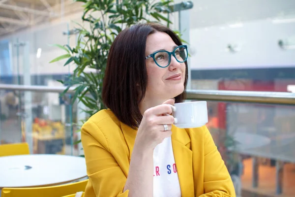Koffiepauze. Brunette meisje in een gele jas met bril, koffie drinken aan een witte tafel. Rusten na het winkelen. — Stockfoto