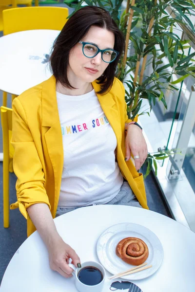 Koffiepauze. Brunette meisje in een gele jas met bril, koffie drinken aan een witte tafel. Rusten na het winkelen. — Stockfoto