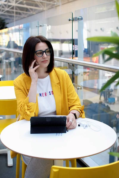 Mooie schattige brunette in een gele jas praten op de telefoon. Jonge zakenvrouw in een café met behulp van een digitale Tablet running. Freelance concept — Stockfoto