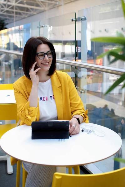 Mooie schattige brunette in een gele jas praten op de telefoon. Jonge zakenvrouw in een café met behulp van een digitale Tablet running. Freelance concept — Stockfoto