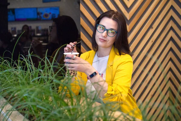 Het meisje houdt een papieren beker met luipaard print en koffie met melk. Koffiepauze. Een brunette in een gele jas en glazen. Direct in de camera kijken — Stockfoto