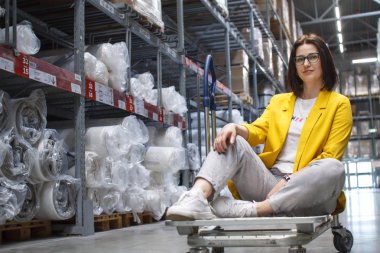 Girl with glasses sitting on a shopping cart in a store in the warehouse. Wholesale and purchase. Self-service warehouse. Choice of goods clipart