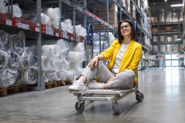 Girl with glasses sitting on a shopping cart in a store in the warehouse. Wholesale and purchase. Self-service warehouse. Choice of goods clipart