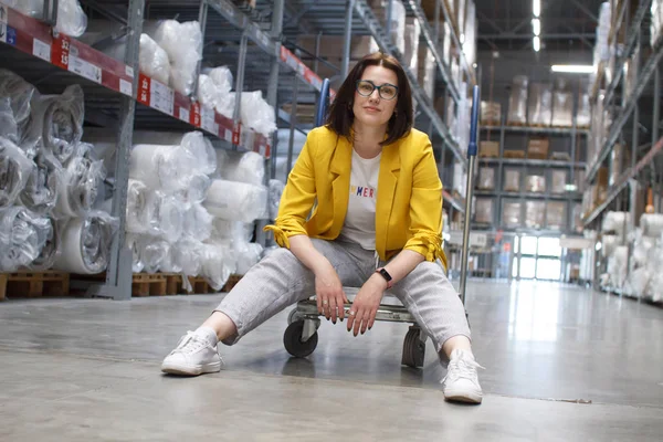 Chica con gafas sentada en un carrito de la compra en una tienda en el almacén. Venta al por mayor y compra. Almacén de autoservicio. Elección de las mercancías — Foto de Stock