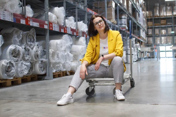 Meisje met bril zittend op een winkelwagen in een winkel in het magazijn. Groothandel en aankoop. Self-service magazijn. Keuze van de goederen — Stockfoto