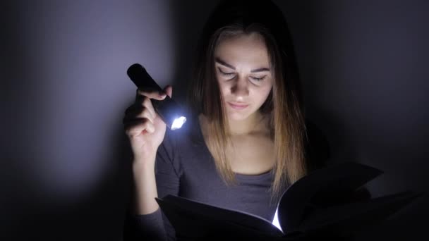 Κοντινό πορτρέτο του έξυπνου κοριτσιού κάθεται με ένα φακό και μετατρέπει τις σελίδες ενός βιβλίου σε ένα σκοτεινό δωμάτιο. 4K. — Αρχείο Βίντεο