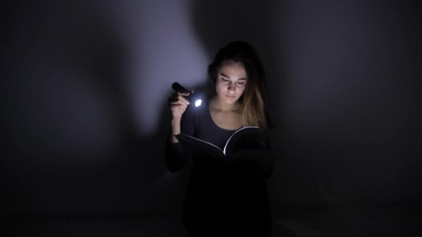 聪明的女孩拿着手电筒坐在黑暗的房间里翻书。4k. — 图库视频影像