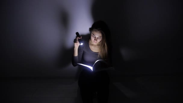 Művelt lány ül egy zseblámpát, és fordul az oldalakat a könyv egy sötét szobában. 4k. — Stock videók