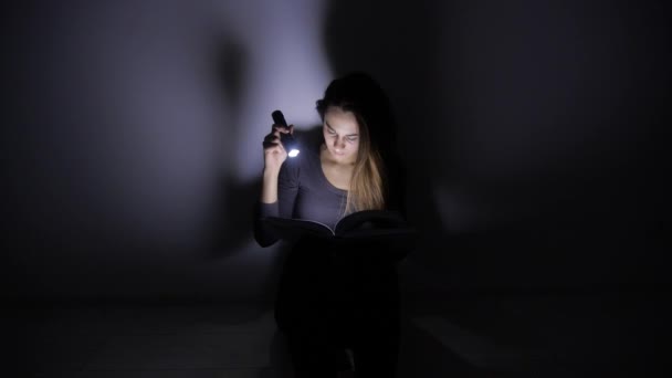 Smart tjej sitter med en ficklampa och läser och förvandlar sidorna i en bok i ett mörkt rum. 4k. — Stockvideo