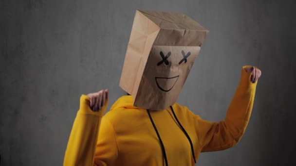 Una chica en sudadera amarilla con bolsas de papel en la cabeza, con un emoticono pintado, sonrisa. Ella está bailando. 4k — Vídeos de Stock