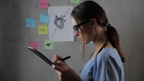 Retrato de una hermosa estudiante de medicina con gafas preparando una presentación, sobre el fondo. Mujer joven dibuja y notas en papel. Concepto: ideas, ciencia, profesor, memoria, biología , — Vídeo de stock