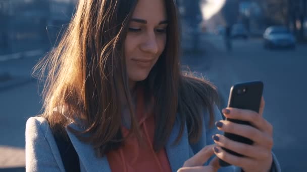 Крупним планом портрет молодої жінки, що йде сонячними вулицями міста, радісна дівчина використовує мобільний телефон, фон заходу сонця . — стокове відео