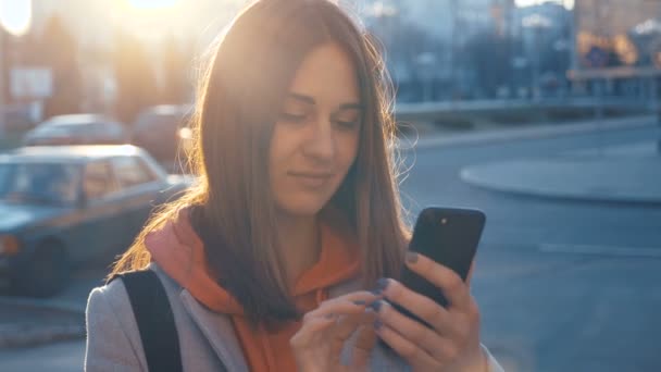 Atractiva joven mujer caminando por las calles soleadas de la ciudad y charlando con amigos, utilizando el teléfono celular al aire libre, fondo de la puesta del sol — Vídeo de stock